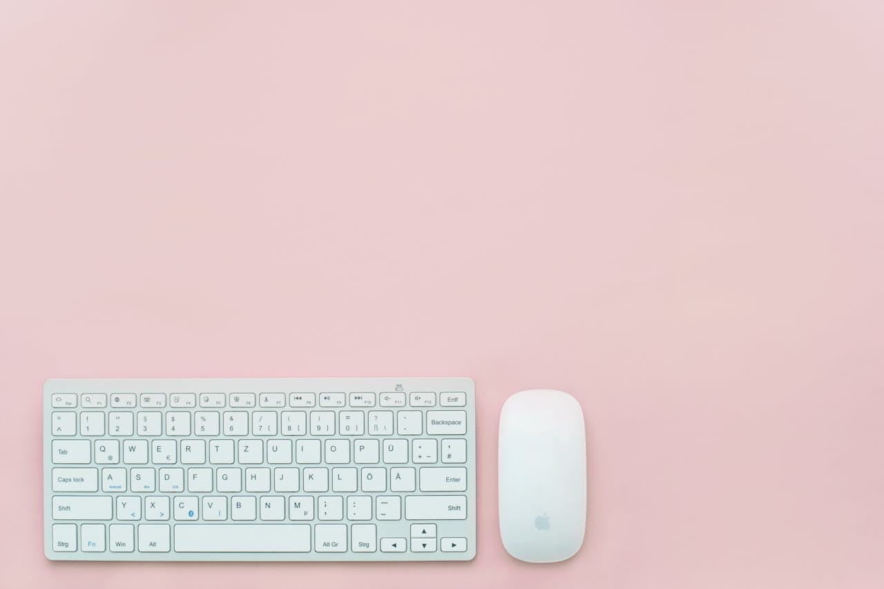 A clean and minimalist flat lay of a white keyboard and mouse on a pastel pink background. Perfect for feminine office themes.